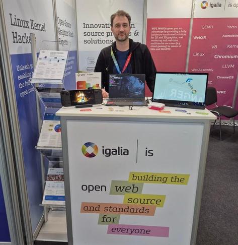 Andrés Gómez at the Igalia booth in Embedded World 2025. In front of him, a tall table with some demos that include a Steam Deck, an Arm laptop and a Raspberry Pi.
