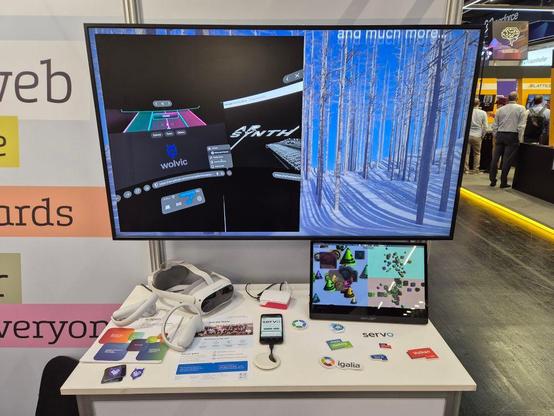 Another demo table at the Igalia booth in Embedded World 2025, with a large screen just behind. The screen shows a demo of Wolvic, a web browser designed for VR devices. On the table, some VR glasses, Servo running on a mobile phone and a Raspberry Pi among others. Also on the table, multiple groups of stickers ready to pick up.