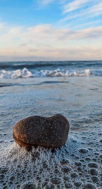 Blick Bodennah auf die Ostsee.  Im Vordergrund ein braune Stein der wie ein Herz geformt ist.  Um ihn aufgeschäumtes flaches Wasser.  Im Hintergrund kommt eine Welle auf uns zu. Blauer Himmel mit kleinen Schleierwolken in zarten rosa.