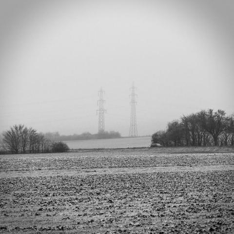 Schwarzweißfoto eines reifbedeckten Feldes im Winter. In der mittleren Distanz befindet sich Gebüsch entlang einer Landstraße, im Hintergrund verschwinden Strommasten halb im Nebel