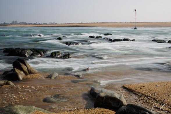 Photo en couleur présentant le flot de la marée descendante à l'embouchure de la rivière d'Etel. De bas en haut, on peut voir le sable de la rive où se trouve le photographe avec quelques rochers, puis la rivière dont on devine la vitesse du courant descendant du fait de la durée d'ouverture de l'obturateur relativement longue, l'eau cascadant sur les quelques rochers maintenant émergés et butant sur la base d'une perche de signalisation. Tout en haut de la photo, on peut voir le sable de la rive opposé sous un ciel encore légèrement brumeux.