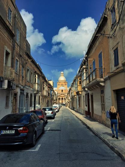 A woman walks up a narrow street of houses in the Maltese city of Paola, in the distance is the dome of Christ The King church.