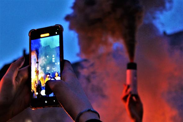 A l'arrière plan, un fumigène noir et orange, au premier plan, le téléphone d'une personne en train de prendre en photo le fumigène