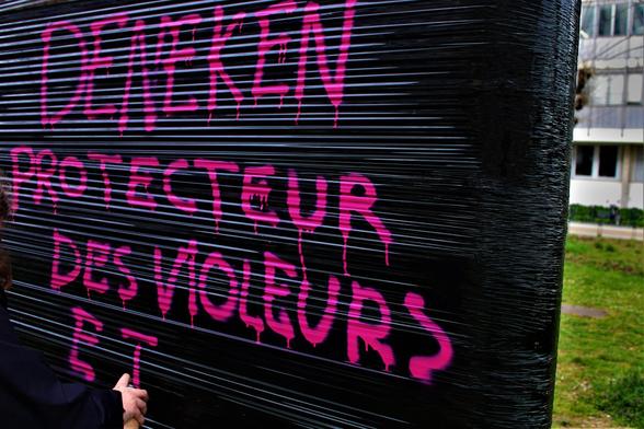 Un•e bloqueur•euse en train de taguer "Deneken protecteur des violeurs" sur une bande de plastique noir accrochée entre deux arbres