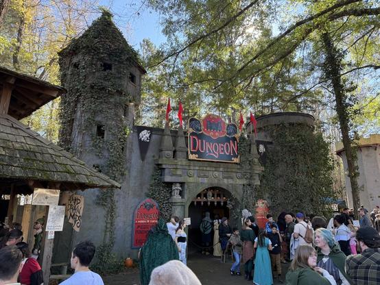 A medieval-themed dungeon attraction at a Renaissance fair, with ivy-covered towers, costumed visitors, and a crowd waiting in line.