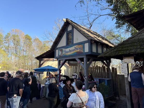 A Renaissance fair hair braiding booth with a medieval-style design, costumed visitors, and a lively crowd waiting in line.