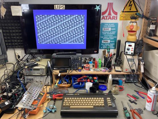 A yellowed Commodore 16 on my workbench with tools scattered around the bench. The monitor shows the output of a little BASIC program, writing "JAN BETA WAS HERE!“ on the screen in an endless loop.