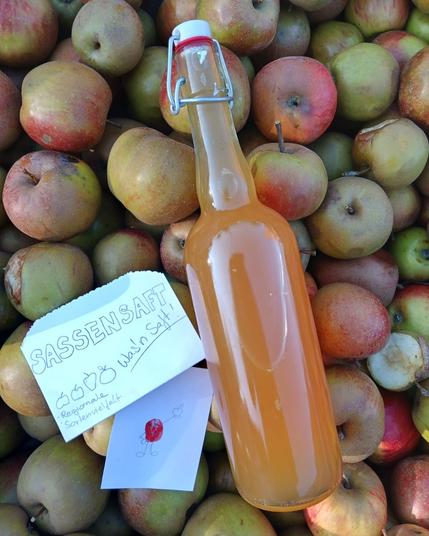 Eine Bügelflasche Apfelsaft liegt auf einem Haufen Äpfel. Auf einem Schild steht: Sassensaft - Was'n Saft!