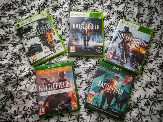 Zdjęcie przedstawia pięć pudełek z grami z serii Battlefield na konsole Xbox 360 i Xbox One. Gry są ułożone na tle wzorzystej tkaniny w czarno-białe liście. Widoczne tytuły to:

Battlefield: Bad Company 2 (Xbox 360),

Battlefield 3 (Xbox 360),

Battlefield 4 (Xbox 360),

Battlefield 1 Rewolucja (Xbox One),

Battlefield 2042 (Xbox One).

Każde pudełko ma charakterystyczną grafikę okładki, prezentującą żołnierzy w dynamicznych scenach wojennych.