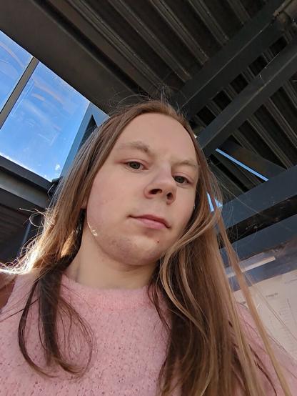 The image captures a woman standing underneath a metallic structure.

The location is a bus station with multiple windows visible in the background.

She is seen from a low angle, looking down at the camera.