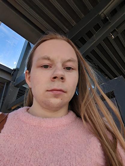 The image captures a woman standing underneath a metallic structure.

The location is a bus station with multiple windows visible in the background.

She is seen from a low angle, looking down at the camera.