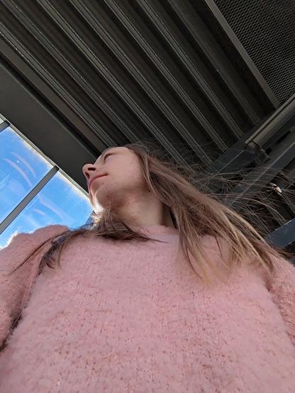 The image captures a woman standing underneath a metallic structure.

The location is a bus station with multiple windows visible in the background.

She is seen from a low angle, looking down at the camera.