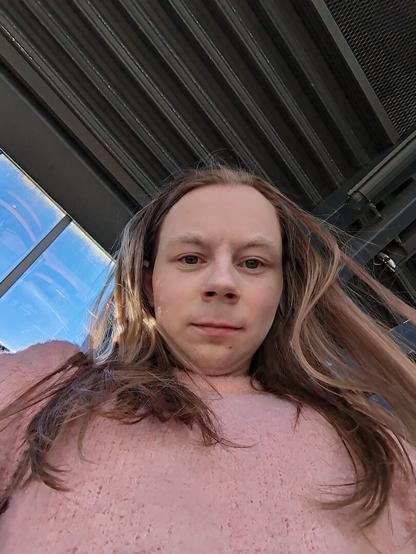 The image captures a woman standing underneath a metallic structure.

The location is a bus station with multiple windows visible in the background.

She is seen from a low angle, looking down at the camera.