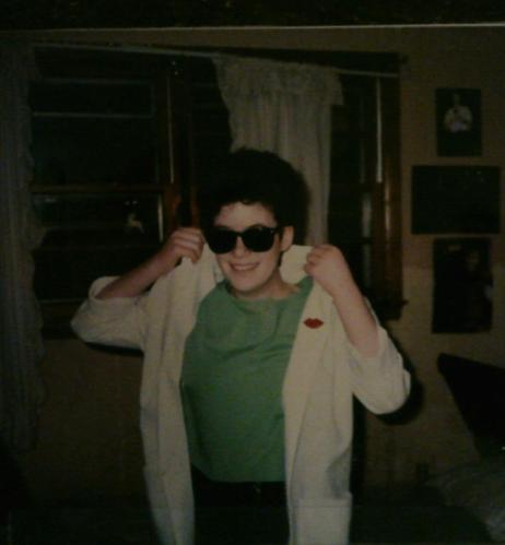 White teenaged girl with short curly hair wears dark sunglasses, a green shirt, and black pants. She's popping the collar on a white single-breasted blazer to which she attached a pin in the shape of red lips. The wall behind her has peeled wallpaper, and the curtain rod is askew. A neatly made bed with collapsed headboard is behind her. Duran Duran posters dot the wall.