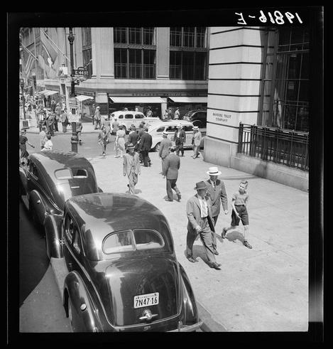 The image is a black and white photograph depicting a bustling city street scene from an elevated perspective, likely taken in New York City. It appears to be captured during the mid-20th century judging by the style of clothing worn by people, vintage cars on the road, and architectural styles.

In the foreground, there are several vehicles parked along the curb: two older model cars with prominent grilles and rounded body shapes indicative of late 1930s or early '40s design. The license plate visible reads "7N4T-16." Pedestrians are walking on a wide sidewalk; some dressed in business attire, suggesting it is possibly during working hours.

Two men dominate the central part of the image: one wearing a fedora hat and dark coat seems to be guiding or escorting another man who appears older. They both exhibit focused expressions as they navigate down the street. Behind them, other people are walking on the sidewalk in various directions; some appear engaged with conversation while others seem more absorbed in their individual paths.

On the right side of the image stands a building labeled "Bankers Trust Company," suggesting an important financial institution is nearby or within view. The surrounding architecture features large commercial buildings with tall windows and signage, typical of urban business districts during that period.

The overall atmosphere conveys movement and activity characteristic of New York City's d [...]