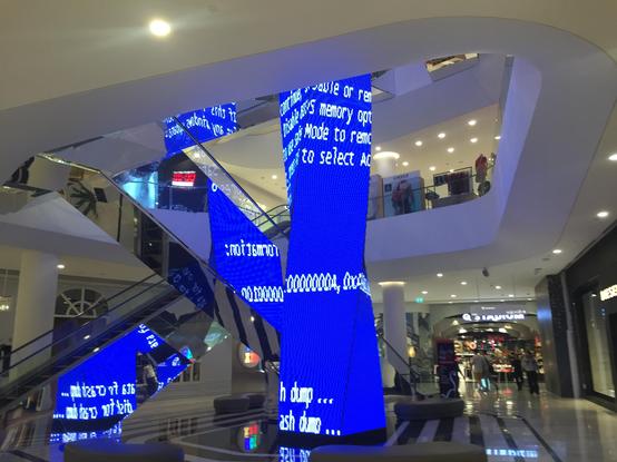 A four-sided twisted pillar located in a morning-empty shopping mall. The pillar is covered with a computer screen to show ads on it. There are two escalators beside it, mounted in opposing directions so they form an X from our point of view. Their sides and undersides are covered in mirrors. The ad-pillar is reflected in those mirrors. When the photographer took the photos, the pillar's screen showed the infamous Microsoft Blue Screen of Death (a full-screen error message, in white text on a blue background).