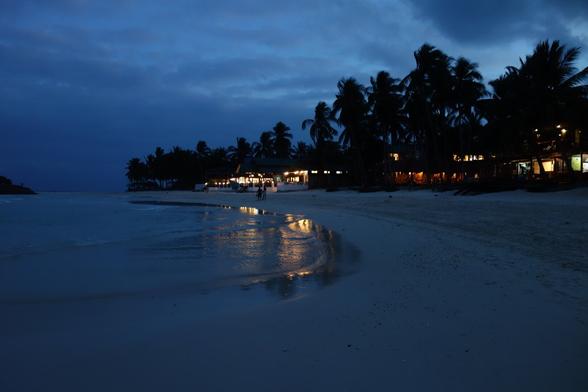 A dusky twilight view of a sandy beach with gentle waves reflecting dim light. Palm trees line the shore, and a building with warm lights is visible in the distance. The sky is a deep blue, suggesting early evening.