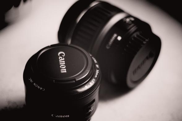 A stylish, monochrome close-up featuring two Canon lenses: prominently highlighting the iconic Canon EF 50mm f/1.8 lens, alongside another softly blurred lens in the background. The image has a minimalistic, cinematic feel with smooth shadows and gentle highlights, emphasizing the sleek contours and clean design of photography gear. Ideal for lovers of photography aesthetics and vintage-inspired gear visuals.