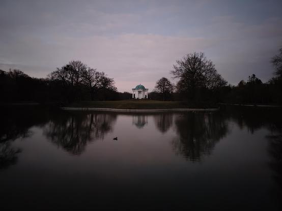 Stiller See um eine kleine Insel in der Mitte mit einem kleine Templchen in der Mitte. Kahle Bäume im Hintergrund und ein Himmel in grau blau mit rosa Farben in leichten Schleierwolken