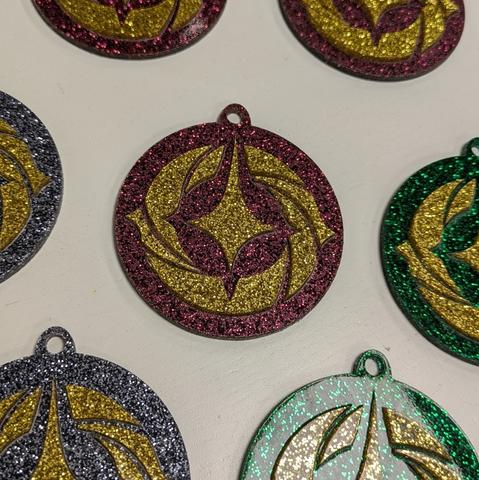 Das Lorcana-Logo als Christbaumschmuck: in der Bildmitte ist das Logo aus gold-glitzerndem Acryl ausgelasert und auf eine rot-glitzernde, runde Grundplatte aufgebracht. Außenherum sind weitere Christbaum-Ornamente in anderen Farben.