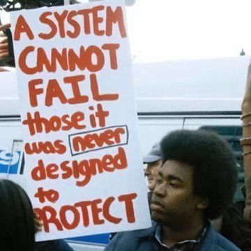 Sign at a demonstration that reads in red “A system cannot fail that was never designed to protect it”