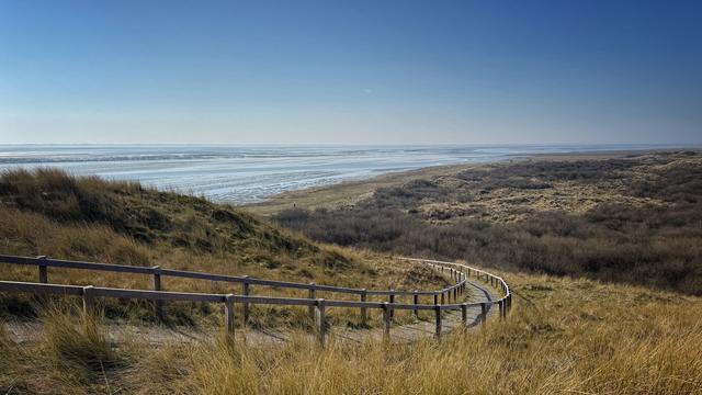 A wide-angle view from a vantage point overlooking the Wadden Sea and the dune landscape of the island of Ameland. A winding path, secured with a wooden railing, leads from the viewpoint to the Wadden Sea. The weather is sunny, bright and friendly with a blue sky.
