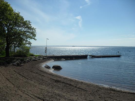 Das Bild zeigt eine ruhige Uferlandschaft mit einem Sandstrand, der sanft ins Wasser übergeht. Der Strand ist von einem Steinhaufen begrenzt, der die kleine Bucht schützt. Im Vordergrund sind einige größere Felsen im flachen Wasser zu sehen.

Ein Steg aus Beton ragt ins Wasser hinein, daneben befindet sich ein schmaler Holzsteg mit einer Leiter am Ende, die ins Wasser führt. Eine Laterne mit einem Rettungsring ist am Rand des Stegs befestigt.

Links im Bild steht ein großer, grüner Baum, dessen Äste sich in Richtung Wasser neigen. Der Himmel ist klar und blau mit wenigen, feinen Wolken. Die Sonne spiegelt sich auf der Wasseroberfläche, wodurch ein funkelndes Glitzern entsteht. Am Horizont ist ein kleines Segelschiff erkennbar. Die gesamte Szene wirkt friedlich und einladend.