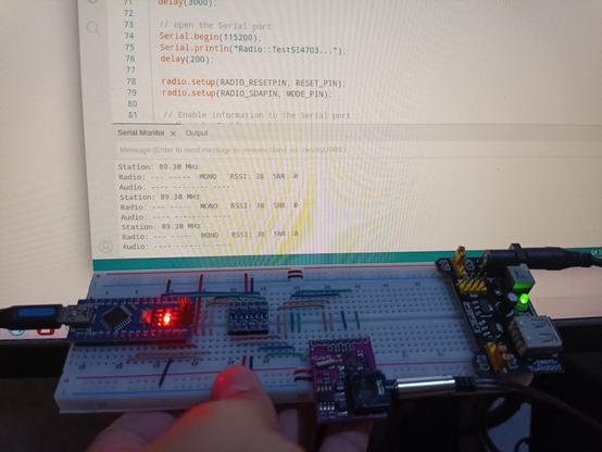 Me holding the breakout board with Arduino Nano clone, a logic level converter, the Si4703 breakout board, and external power supply, everything is powered on. Behind the breadboard is the computer monitor, showing Arduino IDE with serial monitor printing logs that shows that everything works now.