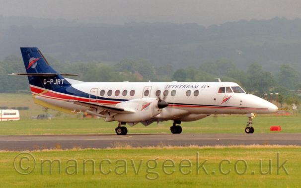 Side view of a white, twin engined jet airliner taxiing along a runway from left to right with flaps extended, suggesting it has just landed.
The plane is almost all white, with a blue, red, and orange stripe running along the body, getting wider as it sweeps up in to the tail.
The tail is mostly blue, with a logo of a white circle with a red and blue lightning flash through it.
The same logo is repeated on the planes nose, only this time the circle is grey.
There are black "Jetstream 41" titles on the upper forward fuselage.
Green grass fills the foreground, with more grass in the background leading up to trees on a hillside in the distance, slowly being lost in haze.
A white ambulance with a red stripe along the body can be seen in the background on the left of the frame.
Grey sky fills the rest of the image.