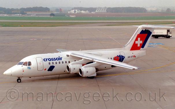 Side view of a white, high-winged, 4 engined jet airliner taxiing from right to left, following a yellow line painted on the concrete floor.
There are blue "CrossAir" titles on the upper forward fuselage, and a small image of a blue flying elephant under the cockpit windows.
There is a blue splotch on the lower forward fuselage, with another blue splotch overlaid with a red splotch on the rear fuselage, and again on the tail, the red splotch here being squared off and having a large white plus sign in the middle.
Concrete apron fills most of the rest of the frame, with grass further back leading up to a grey hangar on the left with planes parked in front, and a rather distinctive large white building in the middle, with trees beyond, under grim grey skies.