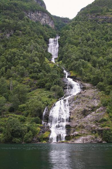 A majestic waterfall cascading down a lush green mountainside in Norway, surrounded by dense forest and reflecting in the calm fjord below.