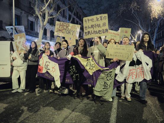 This image shows a group of predominantly Asian women participating in a feminist protest on a city street at night. They are holding handmade signs and banners with messages in multiple languages, including English, Spanish, and Chinese, advocating for gender equality and social justice. The main banner they are holding is a long piece of fabric with a purple base, decorated with women's cloths featuring Chinese characters. The atmosphere is one of solidarity and determination as the group marches together.

Aquest imatge mostra un grup de dones predominantment asiàtiques participant en una protesta feminista en un carrer de la ciutat a la nit. Sostenen cartells i pancartes fetes a mà amb missatges en diversos idiomes, incloent anglès, espanyol i xinès, defensant la igualtat de gènere i la justícia social. La pancarta principal que porten és un tros llarg de tela de base porpra, decorat amb roba de dona amb caràcters xinesos. L’ambient és de solidaritat i determinació mentre el grup marxa junt.