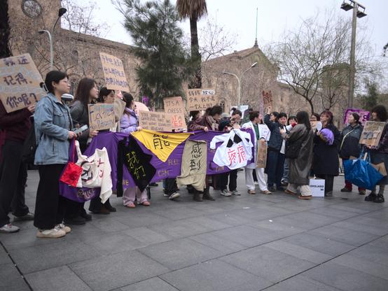 This image, similar to the previous one, shows the protest taking place in an open urban plaza with historic stone buildings and leafless trees in the background. The protesters hold additional signs with new messages, such as “You should be grateful that we just want equality, not revenge” and “It’s a dress, not a yes.” One sign also directly challenges patriarchy with explicit language.

Aquesta imatge, similar a l’anterior, mostra la protesta tenint lloc en una plaça urbana oberta, amb edificis històrics de pedra i arbres sense fulles al fons. Les manifestants sostenen cartells addicionals amb nous missatges, com ara “Hauries d’estar agraït que només volem igualtat, no venjança” i “És un vestit, no un sí.” Un altre cartell també desafia directament el patriarcat amb un llenguatge explícit.
