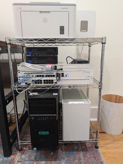 My lab in our new house. On the top is a HP LaserJet printer and a T-Mobile router. In the middle are two Minisforum MS-01 PCs, a CSS610-8G-2S+IN (AP switch), CRS312-4C+8XG-RM (core switch) and CCR2004-16G-2S+ (core router). On the bottom is a HPE ProLiant ML30 Gen9 and a homebuilt i7-13700F PC in a Cooler Master NR200 case.