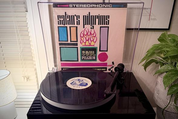 A black vinyl record spins on a black turntable next to a green houseplant. The record sleeve stands up in the turntable’s dust cover behind the platter.