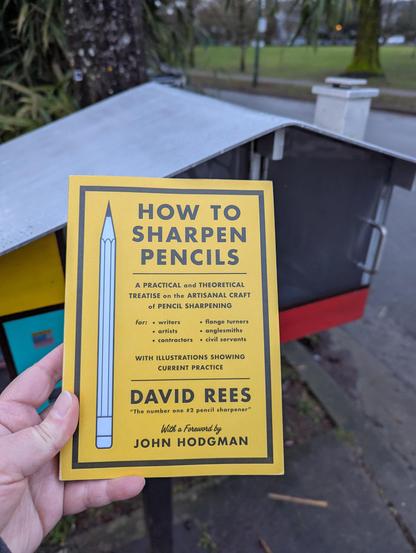 I'm holding a yellow paperback book entitled "How To Sharpen Pencils: A Practical and Theoretical Treatise on the Artisanal Craft of Pencil Sharpening" by David Rees. In the background, there's a little free library. It is a metal and plexiglass house.