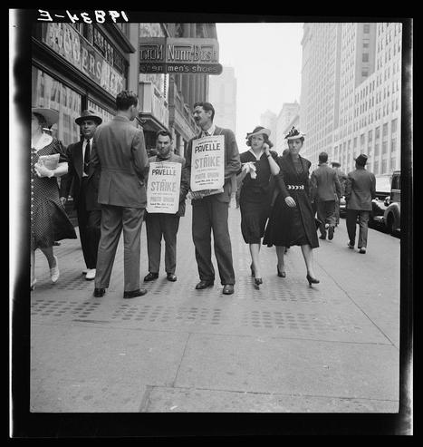 A black and white photograph depicting a group of individuals, likely from the mid-20th century based on fashion style. The scene unfolds on an urban sidewalk with tall buildings in the background indicative of New York City's 1930s or '40s atmosphere. Four people prominently stand holding picket signs bearing phrases related to labor action and union organization.

Two women wear dark, vintage dresses; one holds a small flag, while another clutches papers or a list, possibly for distribution during the strike protest. A man beside them wears a suit and hat with his back turned partially towards the camera, engaged in conversation with an onlooker from across the street.
In front of him are two more individuals: one male holding a sign that reads 'PAVELLE STRIKE PHOTO UNION' alongside another individual whose face is partly obscured. The group's attire indicates they're partaking or supporting this labor action.

The surroundings include pedestrians, suggesting public interest in their protest. Street signs and storefronts, including "Nunn-Bush" shoe store with a visible phone number hint at the commercial nature of Fifth Avenue. It encapsulates both the social struggle during that era amidst everyday life's backdrop.