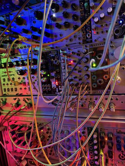 The front of a rainbow lit collection of synth modules connected with lots of cables, there are rows and columns of dials and knobs and switches and jacks everywhere. The different colors of light give it an oily metal appearance.