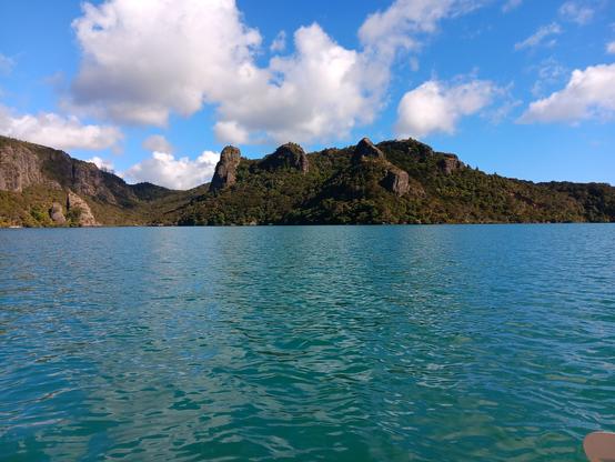 Looking across the water to some tree covered hills and a blue sky w puffy cloud beyond. The hills have interesting rocky lumps.