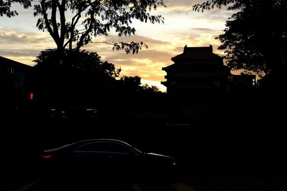View of a sunset sky framed by the silhouettes of a temple and trees, with the silhouette of a car in the lower left part of the picture.