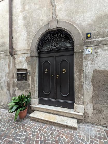 A straight-on shot of a dark, double-doored entrance set within a weathered, stone-textured wall. The doorway features an arched top with decorative wrought iron detailing above the doors. Two small stone steps lead up to the entrance. To the left is a small plant in a terracotta pot, and a letterbox in the wall. A small number plaque reading "30" is visible on the right. It is decorated with lemons. Another plaque just above the number plaque also shows a lemon. The ground is paved with cobblestones.