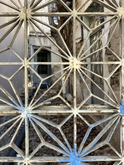 View through the wrought iron garden door of an old Athenian vernacular house, star-patterned and once white, now touched with blue spray paint. The narrow courtyard is out of focus and covered in rubble. A low outbuilding in the back has an open door. The left wall is blank, on the right, the main house, a tree growing too near the wall. There’s another wrought iron door in a different pattern set into that wall.