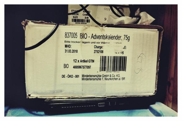 Farbfoto eines weißen Kartons mit einem Aufkleber, der besagt, dass darin BIO-Adventskalender mit einem Gewicht von 75 g enthalten sind. Wird mittlerweile jedoch zur Lagerung von Fotos in Taschen verwendet.