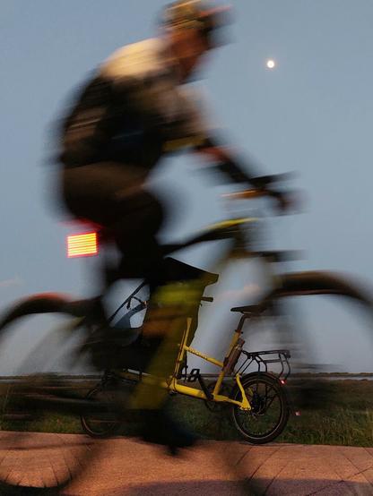 Ciclista desfocado  e por trás uma bicicleta front loader pequena amarela m. Fim do dia com a lua a aparecer. Cabeça do ciclista em linha com a lua