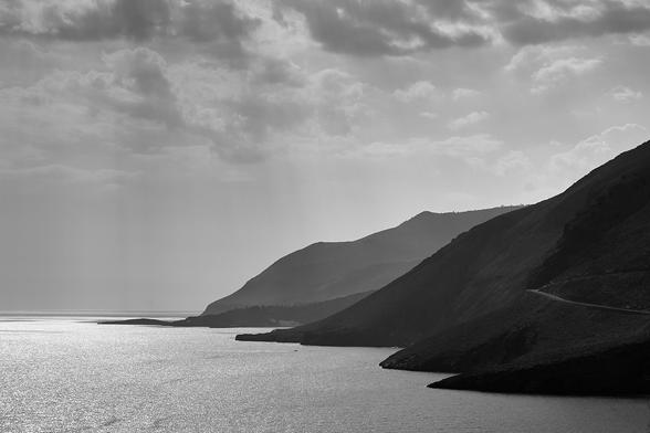 The monochrome photo displays a coastline, with the mountainous coast on the right and the sea to the left. Through the cloudy sky coming sunlight is reflected by the sea.