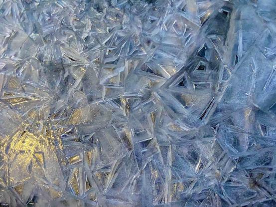 Thick sidewalk ice that has cracked into squares and triangles and sometimes even graffiti-looking letters. Most of is it white and blueish, and in the lower left is reflected some yellow streetlight