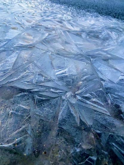 More cracked sidewalk ice, seen from closer to the ground, in a diagonal swath across the frame, with clear black pavement at the upper right.