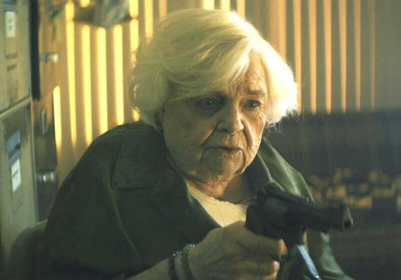 June Squibb in Thelma. She is a room with vertical blinds behind her. She wears a dark sweater. Her gray hair is short.