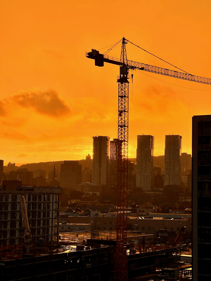 The image depicts a construction scene at sunset. A tall red construction crane stands out against a vibrant orange sky filled with clouds. In the background, several high-rise buildings create silhouettes that contrast with the bright sky. The foreground features a partially constructed building and various construction materials, reflecting the warm colours of the sunset. The overall atmosphere conveys a sense of urban development and the beauty of the sunset.