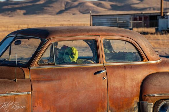 A photograph of a parked old rusted car with three green extraterrestrial aliens sitting inside looking out.